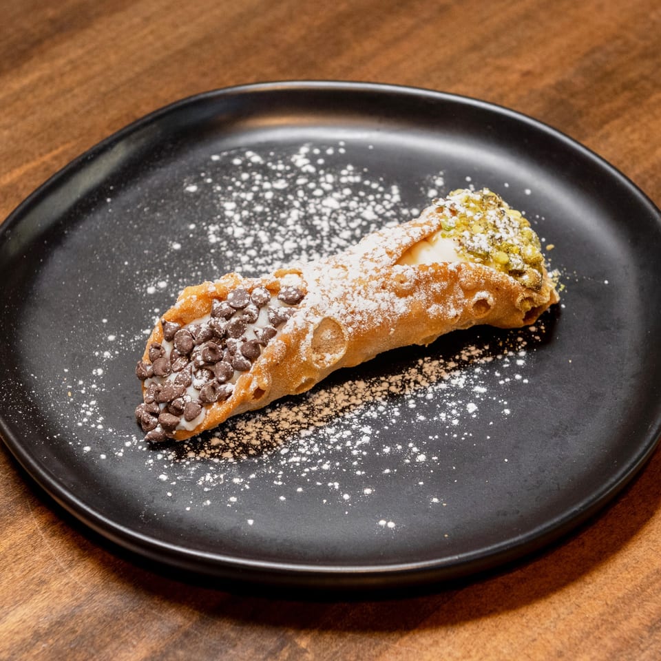 A cannoli from CHAR Pizzeria, dusted with powdered sugar, chocolate chips, and pistachios on a black plate.