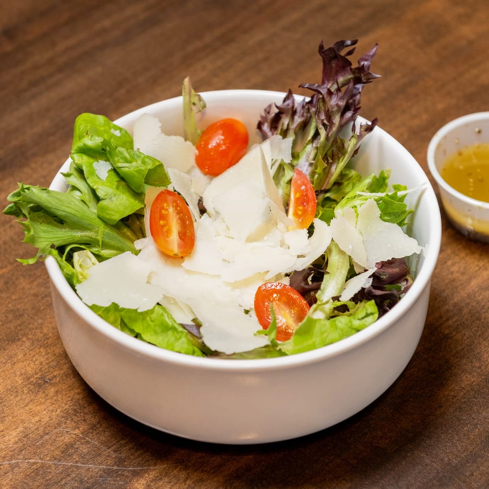 A white bowl of fresh mixed greens, cherry tomatoes, and shaved Parmesan cheese, with dressing on the side.