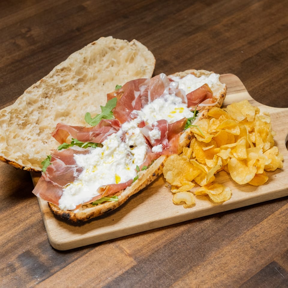 Open-faced sandwich with prosciutto, burrata, arugula, and olive oil, served with potato chips.