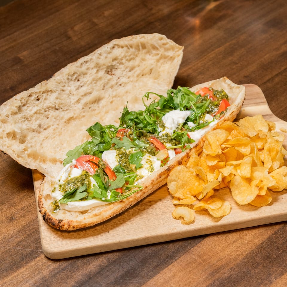 Open-faced sandwich with mozzarella, arugula, tomatoes, pesto, and a side of chips on a wooden board.