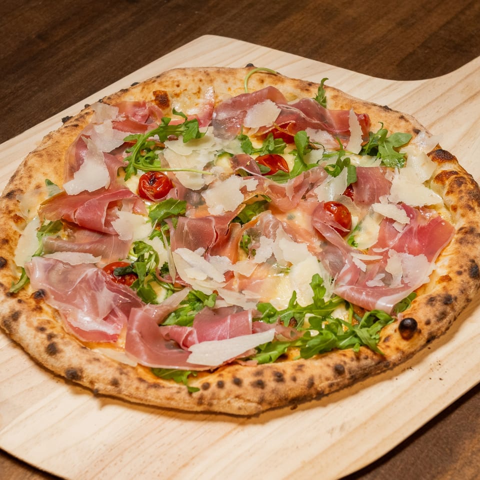 CHAR Pizzeria's pizza with prosciutto, arugula, cherry tomatoes, and shaved parmesan on a wooden peel.