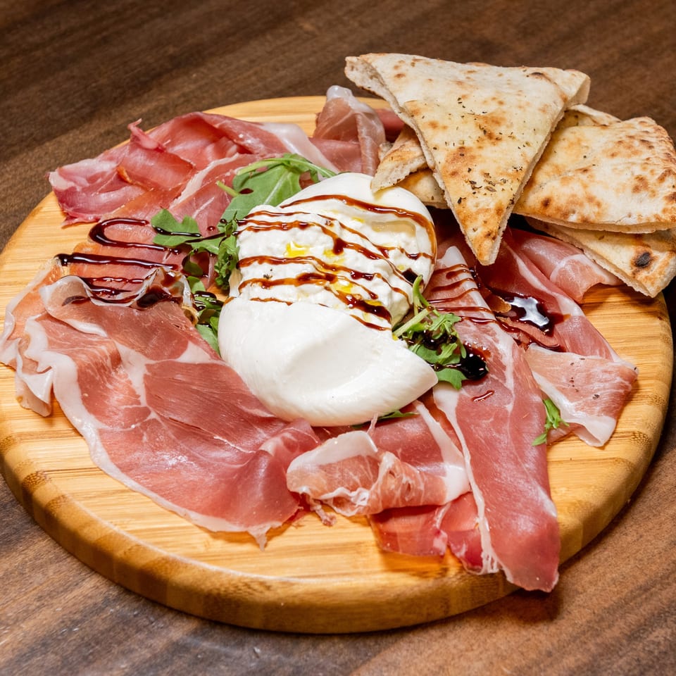 A wooden board with prosciutto, burrata cheese drizzled with balsamic glaze, arugula, and flatbread.